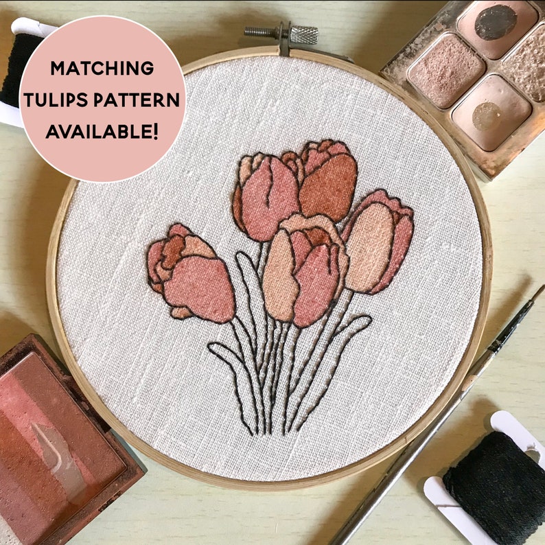 May include: A close-up of a hoop with a white fabric background featuring an embroidered design of a bouquet of pink and peach tulips. The hoop is surrounded by embroidery supplies, including a spool of black thread, a needle, and a small container of blush-colored makeup.