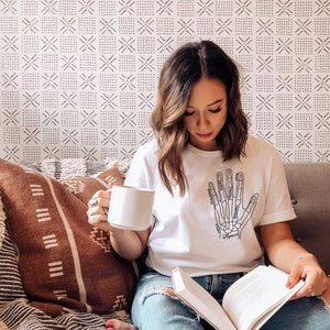 May include: A woman wearing a white t-shirt with a black and white illustration of a hand skeleton. The shirt has the names of the bones labeled. She is sitting on a couch with a book in her lap and a white mug in her hand.