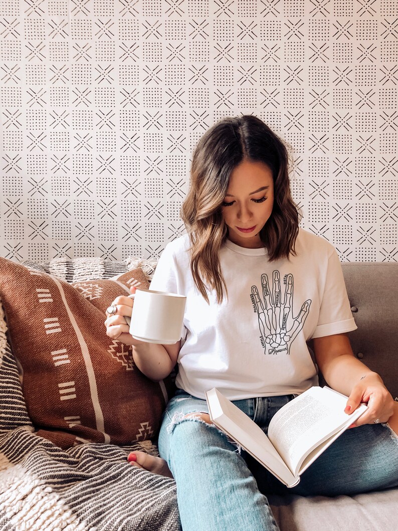 May include: A woman wearing a white t-shirt with a black and white illustration of a hand skeleton. The shirt has the names of the bones labeled. She is sitting on a couch with a book in her lap and a white mug in her hand.