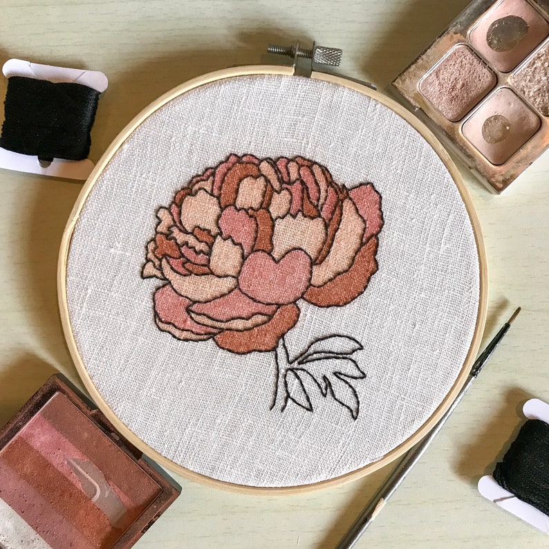 May include: A close-up of a hand-stitched embroidery hoop with a pink and brown peony flower design on a white linen fabric.