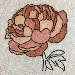 May include: A close-up of a brown and pink embroidered flower with a black outline. The flower is on a white linen background.