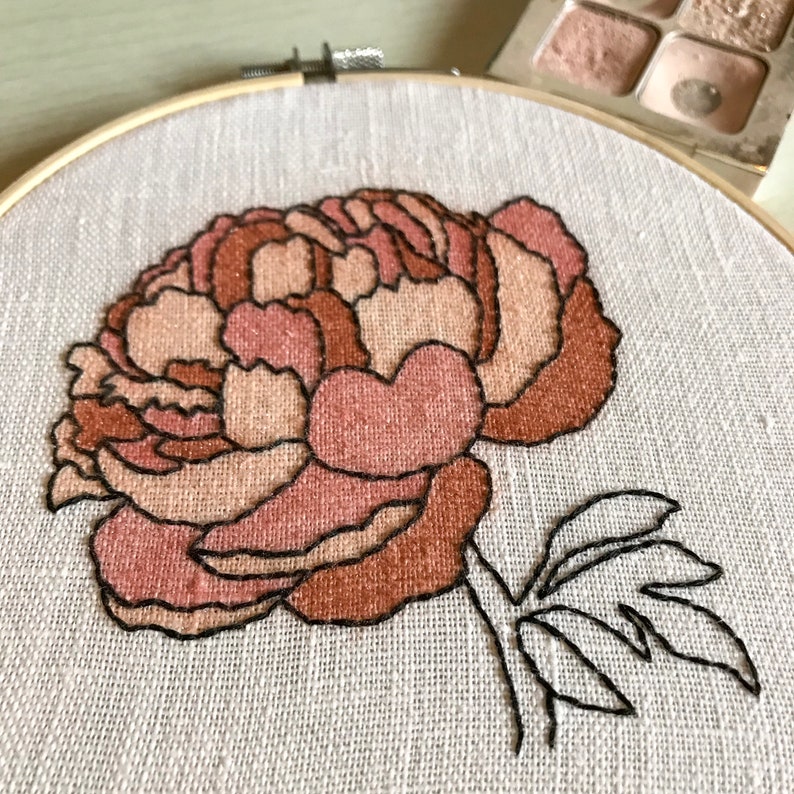 May include: A close-up of a hand-stitched embroidery hoop featuring a large, pink and brown peony flower with a single leaf. The flower is outlined in black thread.