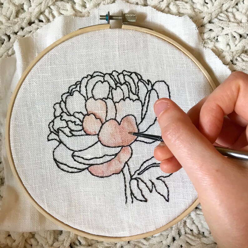 May include: A close-up of a hand painting a pink peony flower on a white fabric stretched in an embroidery hoop. The flower is outlined in black thread.