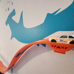 May include: A white toy race car with yellow and red racing stripes and the number 84 printed on the side, positioned on an orange plastic track mounted to a wall. The track curves along the wall, which features a blue hammerhead shark decal.