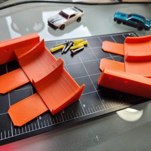 Könnte beinhalten: Zwei orangefarbene Plastikrampen für Spielzeugautos, mit einem schwarzen Gitterhintergrund. Die Rampen sind für die Verwendung mit Spielzeugautos konzipiert und aus strapazierfähigem Kunststoff gefertigt.