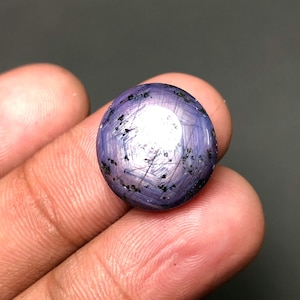 Puede incluir: Una gema redonda y pulida con efecto estrella. La gema es de color púrpura intenso con inclusiones negras y un centro brillante y reflectante. La piedra se sostiene en una mano.