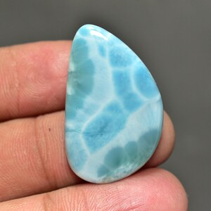 Puede incluir: Una gema pulida en forma de lágrima con un patrón azul claro y blanco. La piedra tiene una superficie lisa y brillante y se sostiene en una mano. El color de la gema se asemeja al océano, con patrones arremolinados de blanco y azul más oscuro.
