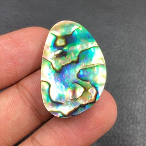 May include: A polished abalone shell gemstone with iridescent colors of green, blue, and gold. The oval-shaped stone has a smooth, glossy surface and a unique, natural pattern. The gemstone is held in a hand.