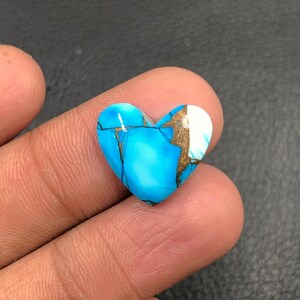 Può includere: Una gemma a forma di cuore con una vibrante tonalità turchese, accentuata da venature dorate e bianche. La pietra lucida è tenuta in una mano, mostrando la sua superficie liscia e le sue uniche variazioni di colore. Ideale per la creazione di gioielli.