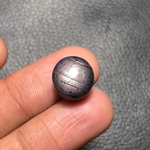 May include: A polished, spherical gemstone in dark blue and silver. The gem has a reflective surface with visible linear patterns and a subtle, speckled texture. The sphere is held between two fingers.