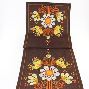 Può includere: Runner da tavolo ricamato marrone con un motivo floreale. Il design presenta grandi fiori bianchi e gialli con centri e accenti arancioni. Il runner ha una forma rettangolare ed è realizzato in tessuto.