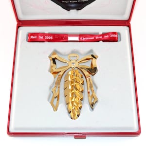 May include: A gold-colored Christmas ornament in a red and white box. The ornament is shaped like a bow and pinecone. The box contains a red ribbon with the text "Noel Jul 2005" and "Christmas Noel 2005".