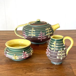 May include: A vintage tea set with a teapot, creamer, and sugar bowl. The set features a green, yellow, and purple color scheme with a striped design. The teapot has a curved handle and spout. The sugar bowl and creamer have matching designs.