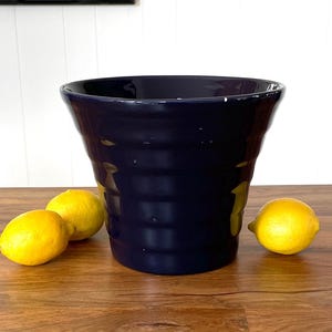 May include: A dark blue ceramic vase with a flared rim and a ribbed design. Three yellow lemons are placed on a wooden table in front of the vase.