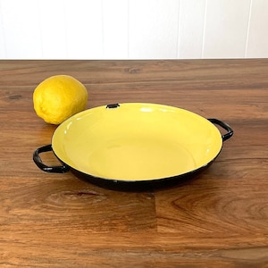 May include: A yellow enamelware baking dish with black handles on a wooden surface. A yellow lemon sits next to the dish.