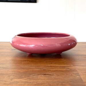 May include: A low, round, pink ceramic bowl with a glossy finish. The bowl sits on a wooden surface. A black framed mirror hangs in the background, reflecting greenery. The bowl's design is simple and modern, suitable for decorative or functional use.