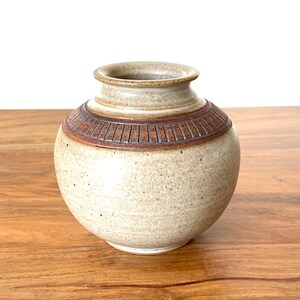 May include: A small, round ceramic vase with a speckled beige finish. The neck of the vase features a decorative band with vertical brown etched lines. The vessel has a wide, bulbous body and a short, flared rim, showcasing a natural, earthy texture.