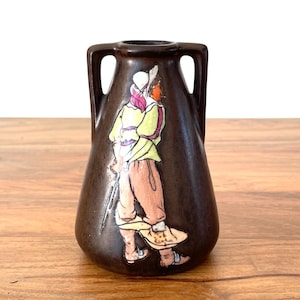 May include: A dark brown ceramic vase with two handles and a painted figure. The figure is wearing a white hat, a yellow jacket, and pink pants. The vase is sitting on a wooden surface.