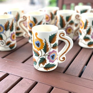 May include: Set of eight white ceramic mugs with floral designs in shades of blue, yellow, orange, and green. The mugs have a handle on each side and are arranged on a wooden table.