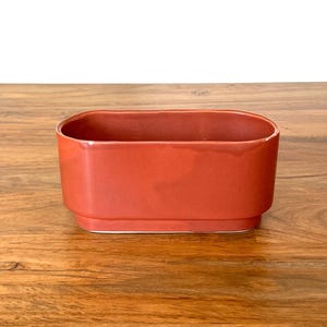 May include: A rectangular, terracotta-colored ceramic planter. The planter has a smooth, matte finish and a simple, modern design. The planter is empty and ready for plants. The planter is sitting on a wooden surface.