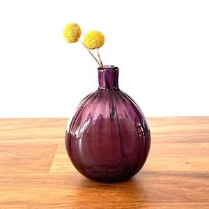 May include: A small, round, deep purple glass vase with vertical ridges, holding two yellow dried flower stems. The vase sits on a wooden surface, with a black framed mirror in the background.