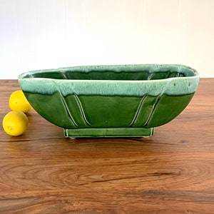 May include: A green ceramic planter with a light green glaze. The planter has a rectangular base and an oval shape. It is sitting on a wooden surface.