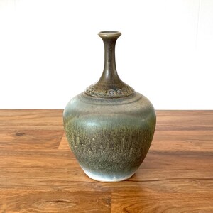 May include: A ceramic vase with a bulbous body and a slender neck. The vase has a gradient of colors, transitioning from a white base to a green-brown midsection and a dark brown top. The vase is displayed on a wooden surface.