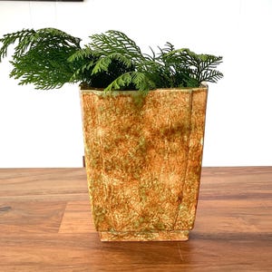 May include: A square, textured planter with a gold and green pattern, filled with green leafy branches. The planter sits on a wooden surface. The background includes a window and white wall.