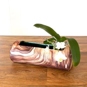 May include: A rectangular, brown and white marbled planter with a green interior, holding an orchid with white flowers and green leaves. The planter sits on a wooden surface, with a window in the background.