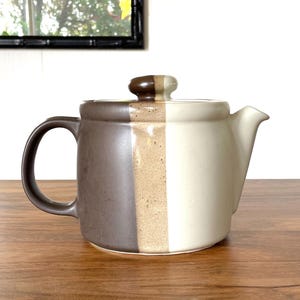 May include: A ceramic teapot with a lid, featuring a color-blocked design. The teapot is divided into three vertical sections: dark brown, beige, and cream. It has a curved handle and a spout. The teapot is sitting on a wooden surface.