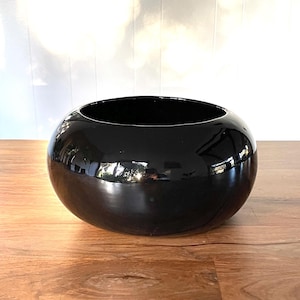 May include: A glossy black, round decorative bowl. The bowl has a smooth, reflective surface and sits on a wooden table. The bowl's shape is a perfect sphere, and it is empty.