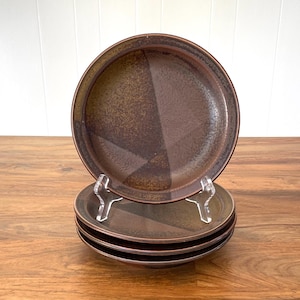 May include: A set of four brown ceramic plates. The plates have a speckled, textured surface with a geometric design. One plate is displayed on a clear stand, with the other three stacked below. The plates are on a wooden surface.