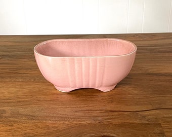May include: A pale pink ceramic planter with a ribbed design. The planter is oval shaped and has a slightly rounded bottom.