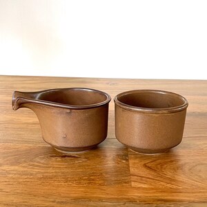 May include: Two small, round, brown ceramic bowls with a matte finish. One bowl features a small, integrated pouring spout on the side, while the other is a simple, open-rimmed circular bowl. Both items have a consistent, earthy brown tone.
