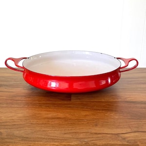 May include: A round, shallow enameled cast iron pan with a vibrant red exterior and a smooth white interior. The pan features two small, curved side handles and a wide, open design, providing a clear view of the contrasting color scheme.