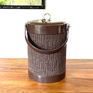 May include: A brown ice bucket with a woven texture and a matching lid. The bucket has a handle and a metal ring on top. The design is vintage and the bucket is sitting on a wooden surface.