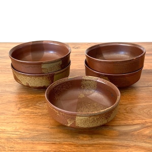 May include: A set of six ceramic bowls. The bowls are a deep brown color with a speckled, golden-brown band around the base. The bowls are stacked in pairs and arranged on a wooden surface.