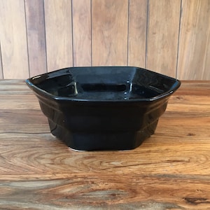 May include: A black octagonal ceramic bowl with a smooth, glossy finish. The bowl is sitting on a wooden surface.