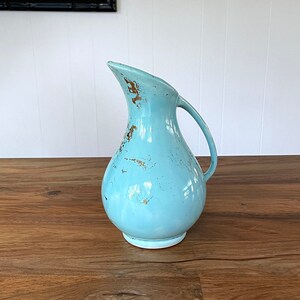May include: A light blue ceramic pitcher with a distressed finish. The pitcher has a rounded body and a curved spout. It is sitting on a wooden table.