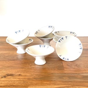 May include: A set of five white ceramic footed bowls, each featuring a series of small, grey triangular patterns along the inner rim. The bowls have a wide, flared opening and a sturdy, tapered base, arranged in a cluster on a wooden surface.