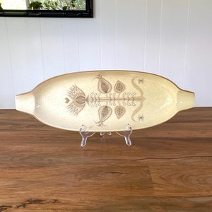 May include: A beige oval serving dish with a brown floral design. The dish is on a wooden surface.