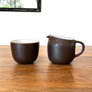 May include: A brown ceramic cup and creamer set with a white interior. The cup is round, and the creamer has a handle and spout. The set is placed on a wooden surface, with a mirror in the background.