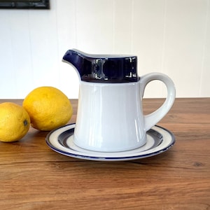 May include: A white ceramic creamer with a blue rim and handle, sitting on a blue and white striped saucer. The creamer is on a wooden table with two lemons.