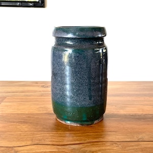 May include: A cylindrical ceramic vase featuring a deep blue and dark green glazed finish. The upper portion of the vase is a mottled dark blue, transitioning into a solid dark green band at the base. It has a simple, straight-sided design.