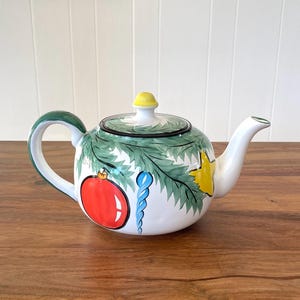 May include: A white ceramic teapot with a Christmas theme. The teapot features a green handle and rim, a yellow lid, and is decorated with a red ornament, blue icicle, and yellow star. The teapot is sitting on a wooden surface.