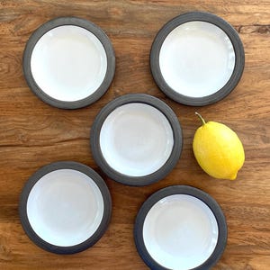 May include: Overhead shot of five small, round, white ceramic dishes with dark gray rims. A bright yellow lemon sits next to the dishes on a wooden surface. The dishes are arranged in a circular pattern.