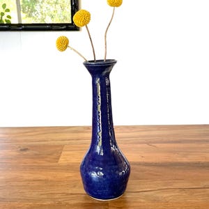 May include: A cobalt blue ceramic vase with a slender neck and a rounded base. The vase holds three yellow globe-shaped flowers. The vase is on a wooden surface, with a window and mirror in the background.