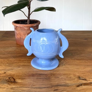 May include: A light blue ceramic vase with two handles and a circular design. The vase is sitting on a wooden table.
