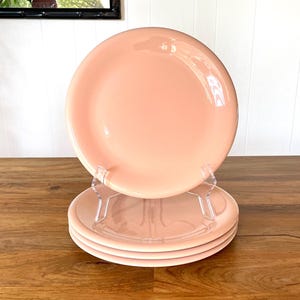 May include: A stack of four round, solid light peach-colored ceramic plates. The top plate is positioned upright on a clear acrylic display stand, while the remaining three plates are stacked horizontally beneath it, creating a uniform set.