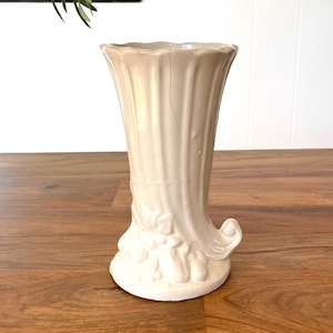 May include: A cream-colored ceramic vase with a fluted design and a decorative base. The vase has a flared top and a curved handle-like detail on the side. The vase is approximately 8 inches tall.